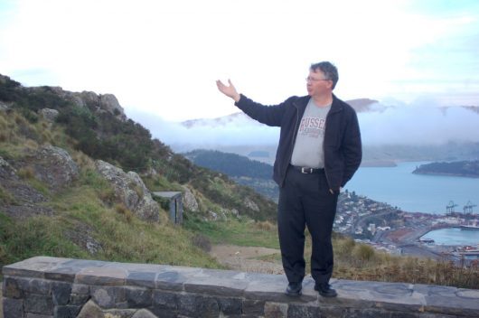 Russell standing on a wall with one arm outstretched as if lecturing on a grand subject, at a scenic spot overlooking Christchurch.