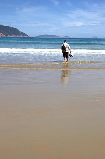Russell from the rear, holding his shoes and walking into the shallows of a beach on a sunny day.