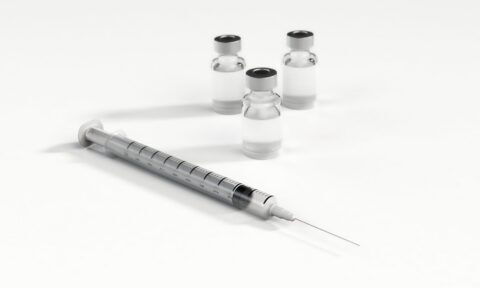 A syringe with a needle is placed in front of three small glass vials containing a clear liquid, all set on a white surface.