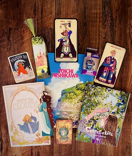 Photos of book covers of Wanderlust by Yoichi Nishkawa, Slices of Life by Qu, and Above the Clouds by Melissa Pagluica. The photo also contains fantasy and brightly colored bookmarks, art cards, and stickers as well as a figurine of Derpy, the blue tiger from Kpop Demon Hunters.