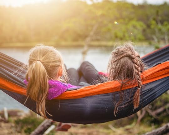 two young girls with brown hair sitting side by side in hammock overlooking a lake. 