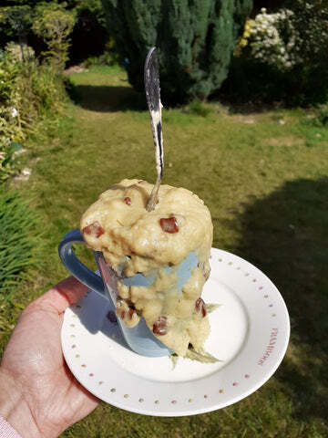 A coffee mug filled to overflowing with a microwave mug cake. I'm holding it up against the background of my sunny garden.