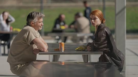 A man in a beige prison uniform and a woman in a dark jacket sit at a picnic table in an outdoor setting, engaging in conversation. Other people are present in the background.