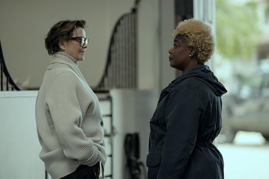 Two women facing each other in profile, one with short hair and glasses wearing a light sweater, and the other with curly blonde hair in a dark coat.