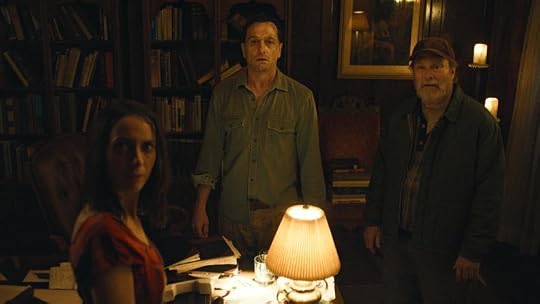 Three people standing in a dimly lit room filled with bookshelves; a woman in a red shirt, a man in a green shirt, and an older man in a jacket, looking surprised or concerned.