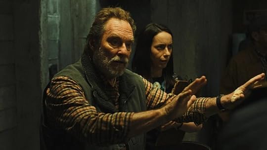 A man with a beard and plaid shirt raises his hands in a gesture of caution or warning, while a woman behind him looks surprised, both standing in a dimly lit room.