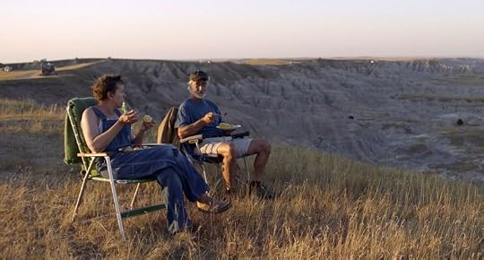 A man and woman sit in camping chairs on a grassy hill, enjoying a meal while overlooking a vast, rugged landscape at sunset.