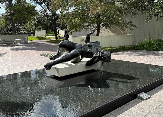 A bronze sculpture of a reclining nude female figure (La Rivière by Aristide Maillol)on a low pedestal in the center of a shallow, dark reflective water pool in an outdoor courtyard.