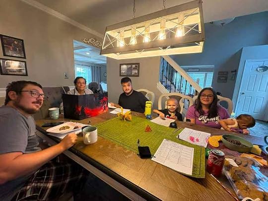 A family gathered around a dining table with various snacks, looking engaged in a board game or role-playing session. The table features a green game mat, colorful dice, and notebooks, while a pendant light hangs above them.