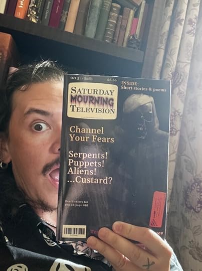 Author Die Booth (a white man with long dark hair, dark eyes and dark moustache) holds up a copy of Saturday Mourning television, which features a shrouded skeleton on the cover which resembles TV listing pages. He has a shocked expression on his face.