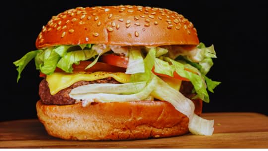 Loaded cheeseburger with melted cheese, tomato, lettuce, and onion spilling out of a sesame seed bun on a wooden board against a black background.