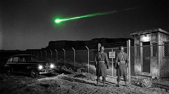Black and white noir photograph depicting the December 1948 green fireball sightings over New Mexico. Two military security guards in period uniforms stand at the chain link perimeter fence of an atomic weapons installation at night, looking upward as a vivid green fireball travels horizontally across the sky above the desert mesa. The fireball is the only color in an otherwise monochrome image. A government sedan idles nearby with headlights illuminated, capturing the moment the atomic security establishment first confronted a phenomenon it could not explain.