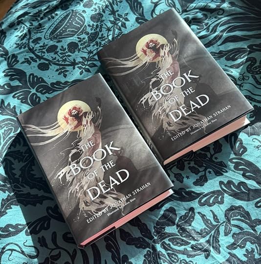 Two copies of a hardcover book like on a teal and black cloth. The covers say THE BOOK OF THE DEAD edited by Jonathan Strahan and illustrated by Galen Dara. The cover has an image of a spooky ghost-like figure with trailing hair and smoke in front of a full moon. It has a skull face and roses in its hair. Perhaps it is a ghost bride. The silhouette of a headstone is in the background.