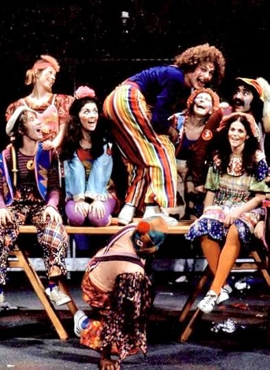 A colorful group of clowns performing on stage, with one clown bending over on a table while others laugh and enjoy the show.