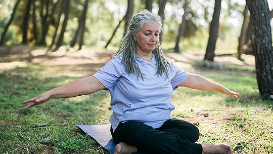 A senior woman with gray hair and a light purple shirt sits cross-legged on a blue yoga mat in a sunny wooded area, with arms stretched out to each side.