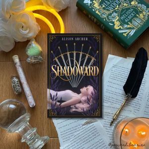 Picture of the book Shadoward on a wooden backdrop surrounded by magical items, including a glowing crescent moon, a crystal ball, a lit candle, and a quill