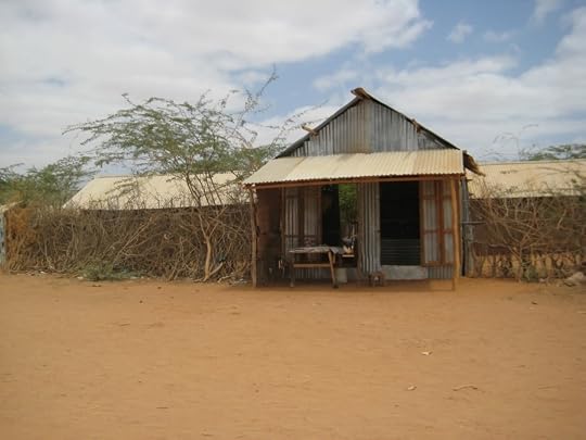 A store with a corrugated metal front, a peaked roof, and a wooden awning over a small table, set in a fence of woven acacia branches. Long, low corrugated-roof shelters stretch behind it on either side, above bare red earth