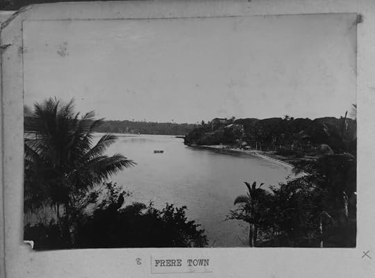 A black-and-white archival photograph, mounted in an album and labeled ���Frere Town,��� looking down through palm trees onto a calm bay. A boat with several people in it crosses the water toward a curved beach backed by dense trees and a few buildings.