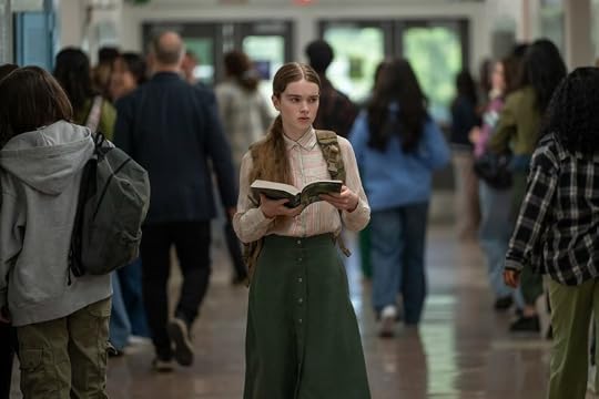 A young girl stands in a busy school hallway, holding a book and looking thoughtfully ahead. She is wearing a striped blouse and a long green skirt, with a backpack slung over one shoulder. Other students pass by her in the background.