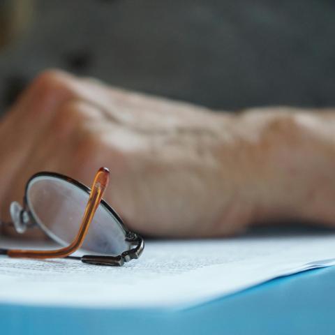 Alt text: On a desk or table, a pair of folded eyeglasses rests on sheets of paper. A person's forearm and hand are lightly resting on the desk behind the glasses.
