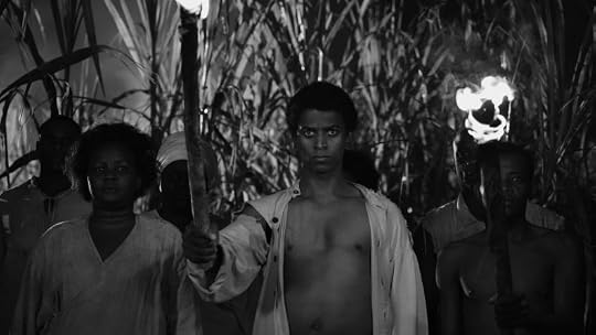A group of individuals holding torches, standing in a field of tall grass, depicted in black and white. The central figure, a young man, has a serious expression and is shirtless, while others are dressed in simple clothing.