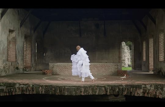 A person in a flowing white outfit is performing a dance on a circular platform within an old, partially ruined building. The walls are made of brick and stone, and the space is dimly lit, featuring some greenery visible through the openings.